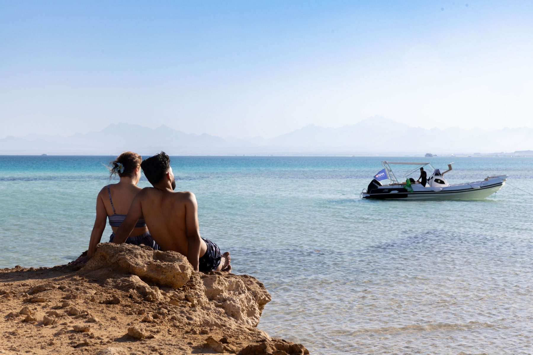 A couple sitting on a quiet Red Sea shoreline while the Maro Redsea speedboat waits in the turquoise shallows
