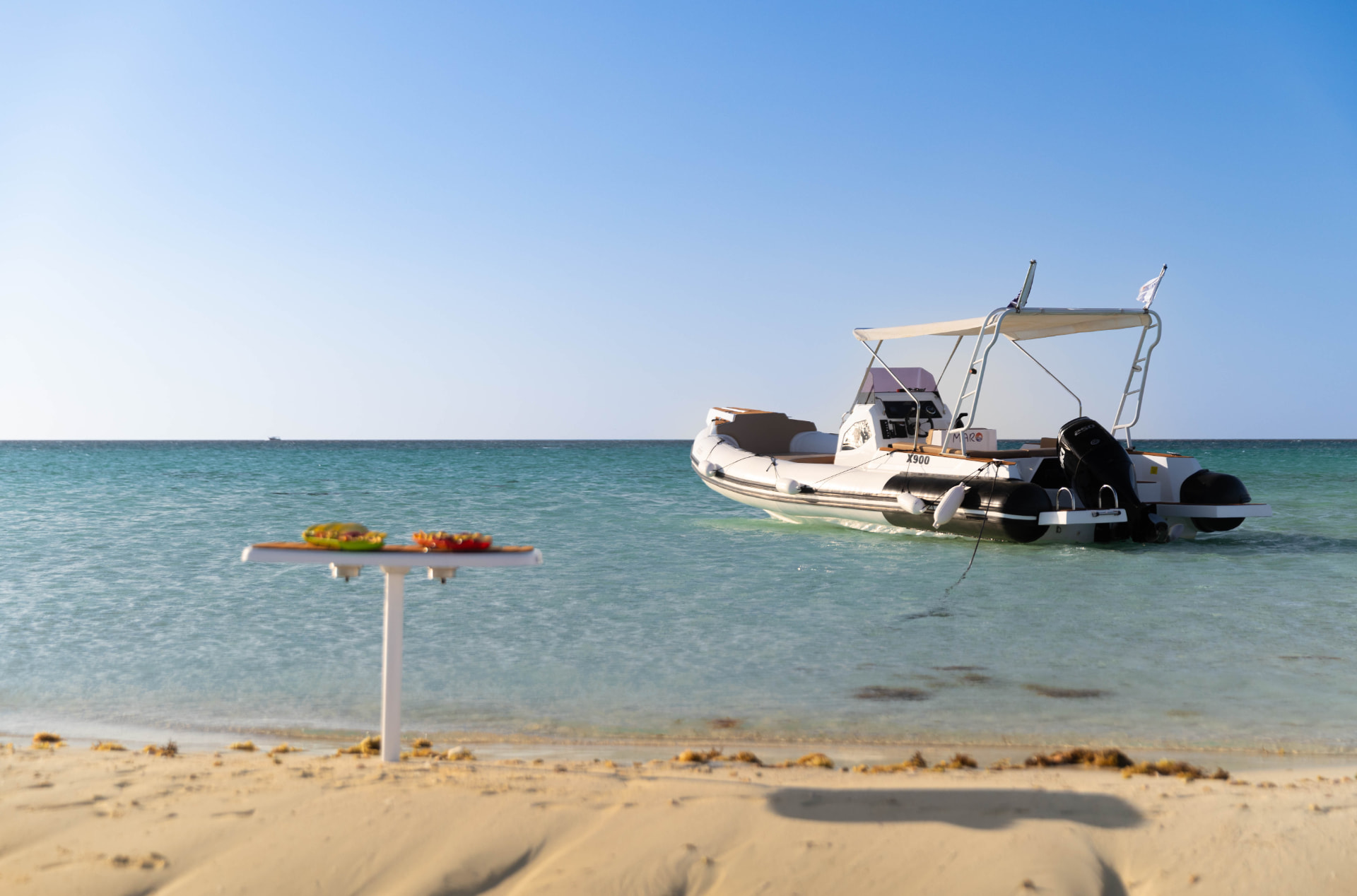 Bateau à moteur MARO X900 ancré dans une eau turquoise peu profonde près d'une plage de sable, avec une table de fruits frais